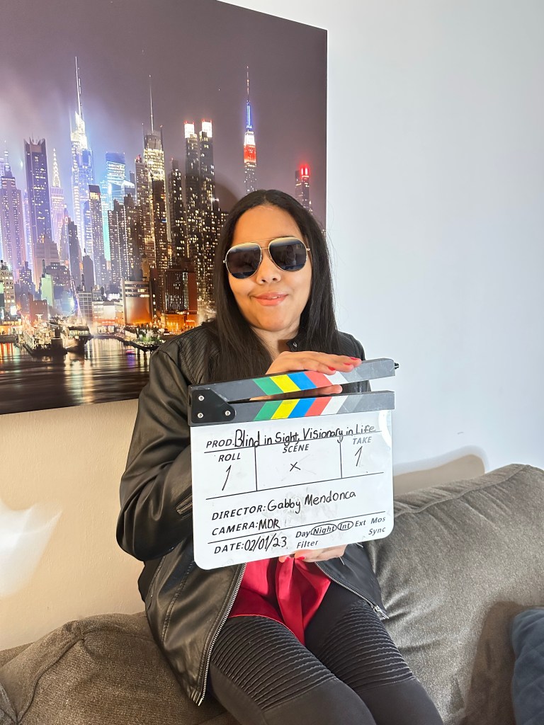 An image of Gabby, a lightskin woman sitting on a couch holding a clapboard that reads: Blind in sight, visionary in life. She is wearing sunglasses and a leather jacket. There is an image of the New York city skyline on the wall behind her.