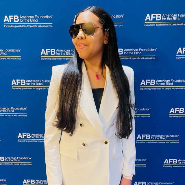 Gabby is wearing a white double breasted jacket paired with black pants and sunglasses, standing in front of an AFB (American Foundation for the Blind) banner. Her long dark hair is draped over both shoulders and she's giving off boss lady vibes.